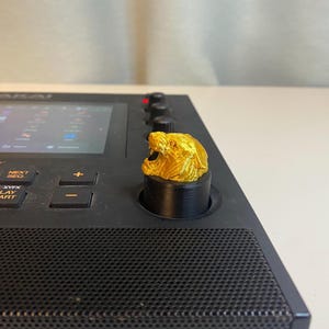 May include: A gold-colored 3D printed knob with a lion's head design, mounted on a black knob for a music production controller.