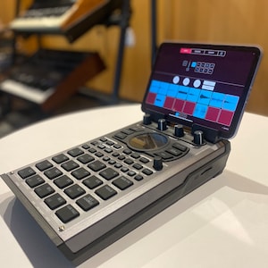 May include: A silver and black music production device with a tablet attached. The tablet screen shows a music production interface with a blue and red colour scheme. The device has a circular knob, multiple buttons, and a grid of pads.