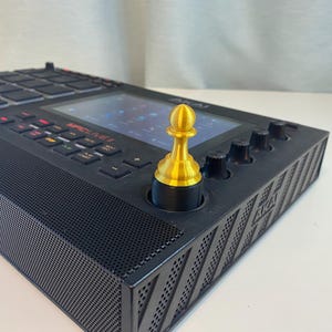 May include: A black music production controller with a gold chess piece on the top knob. The controller has a screen, buttons, and a speaker grill.