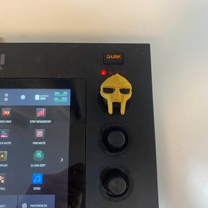 Może przedstawiać: Czarne urządzenie do produkcji muzyki Akai Professional z dołączonym żółtym akcesorium w kształcie maski. Urządzenie posiada ekran dotykowy oraz różne przyciski i pokrętła. Maska jest stylizowaną reprezentacją twarzy.