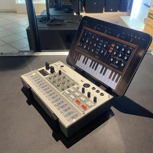 May include: A white and black electronic drum machine with a tablet displaying a synthesizer interface. The tablet is attached to the drum machine with a stand.