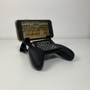 May include: A black phone controller with a phone attached to the top. The phone is displaying a retro video game. The controller has a grid of buttons and two red buttons on the top.