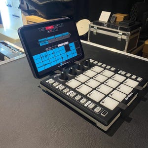 May include: A black and white MIDI controller with 16 pads and knobs, connected to a tablet displaying a music production app.