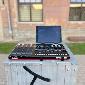 Może przedstawiać: Czarno-czerwony kontroler MIDI Akai MPD32 z tabletem na górze. Kontroler stoi na szarej metalowej powierzchni z czarnym graffiti.