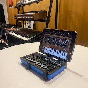 Peut inclure: Un contrôleur MIDI bleu et noir avec une tablette affichant une interface de synthétiseur. La tablette est montée sur le contrôleur à l'aide d'un support.