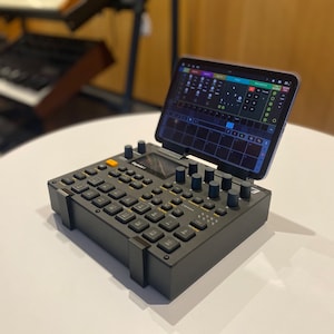 May include: A black music production controller with a tablet attached to the top. The tablet is displaying a music production app with a grid of buttons and sliders. The controller has a variety of buttons, knobs, and pads.