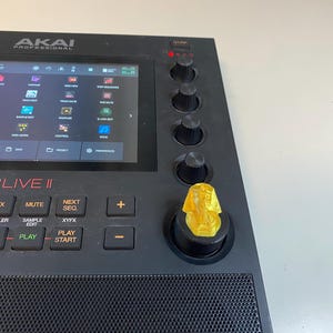 May include: A black Akai MPC Live II music production sampler with a gold pharaoh head knob. The screen displays the menu options for the device. The buttons below the screen include: Main, Mix, Mute, Next Seq, Grid, Sampler, Sample Edit, XYFX, Over Dub, Stop, Play, Play Start.