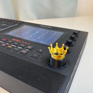 May include: A black Akai MPC Live II music production sampler with a gold crown knob. The screen displays the text "MPC LIVE II".