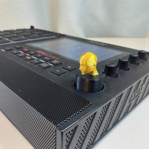 May include: A black music production controller with a gold-colored 3D printed knob. The knob is shaped like a human head. The controller has a screen and a grid of buttons.