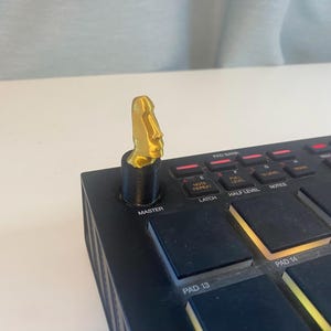 Peut inclure: Un bouton en plastique imprimé en 3D de couleur dorée avec un motif de visage, monté sur un appareil de production musicale électronique noir. L'appareil comporte des boutons étiquetés "Master", "Latch", "Half Level" et "Notes".