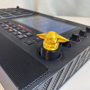 May include: A black music production controller with a gold-colored character figurine on a knob. The controller has a screen, buttons, and a speaker.
