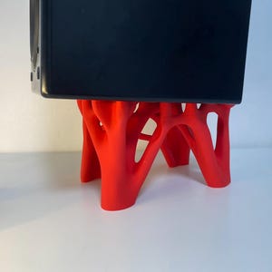 Pode incluir: Um suporte vermelho, impresso em 3D, com um design orgânico único que suporta um objeto retangular preto. A estrutura do suporte assemelha-se a ramos ou raízes entrelaçadas. O suporte foi concebido para segurar um dispositivo.