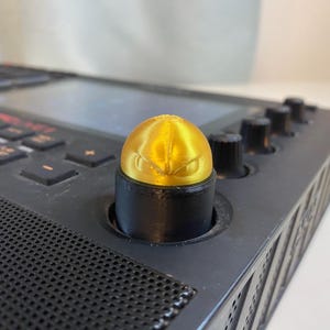 May include: A gold knob with a stylized eye design sits on top of a black knob on a black electronic music production device.