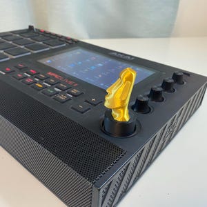 May include: A black Akai MPC Live II music production center with a gold-colored Moai statue knob. The MPC features a screen, buttons, and knobs. The Moai statue is a decorative addition to the music production equipment.