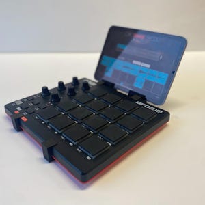 Może przedstawiać: Czarno-czerwony kontroler produkcji muzycznej MPC One z tabletem przymocowanym do górnej części. Tablet wyświetla interfejs produkcji muzycznej.