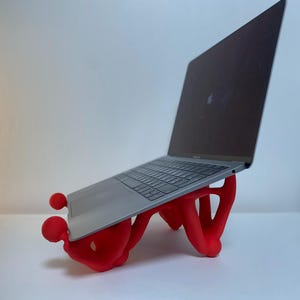 Pode incluir: Um suporte para laptop vermelho, impresso em 3D, com a forma de duas pessoas segurando um laptop. O laptop é prateado e tem um teclado preto.