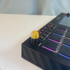 Könnte beinhalten: Ein schwarzer Musikproduktionscontroller mit einem goldenen Knopf. Der Controller hat einen Bildschirm und mehrere Knöpfe und Pads.