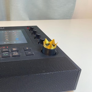 May include: A black music production sampler with a gold crown knob. The sampler has a screen, buttons, and a speaker.