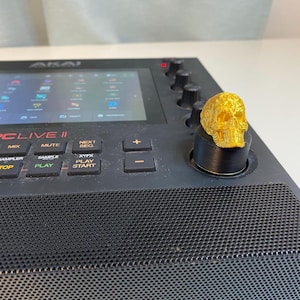 May include: A gold-colored skull knob on a black Akai MPC Live II music production center. The device has a screen, buttons, and knobs. The text "MPC LIVE II" is visible on the device.