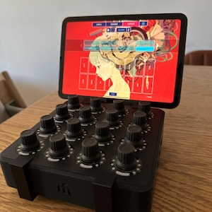 May include: A black music production controller with a grid of knobs and a tablet displaying a colorful interface. The tablet shows a graphic of a woman with a colorful background.