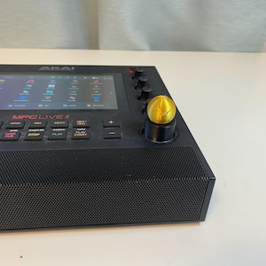 May include: Black Akai MPC Live II music production center with a gold-colored knob. The device has a screen displaying various icons and buttons. The front of the device has a speaker grill and several buttons labeled with functions.