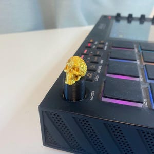 May include: A gold skull knob for a music production controller. The knob is mounted on a black plastic base. The knob is on a black music production controller with a screen and buttons.