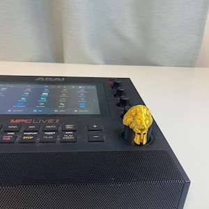 May include: A black Akai MPC Live II music production sampler with a gold lion head knob. The screen displays the MPC Live II interface.