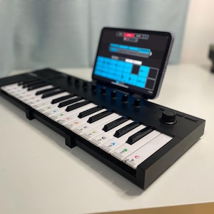 May include: A black MIDI keyboard with white keys and a tablet displaying a music app. The keyboard has a knob and a set of buttons. The keys have colorful labels with musical notes and letters.