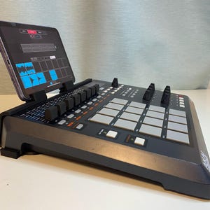 May include: A black electronic music production device with an angled tablet displaying a music production interface. The device has numerous buttons, knobs, and pads. The tablet screen shows a waveform and other controls.