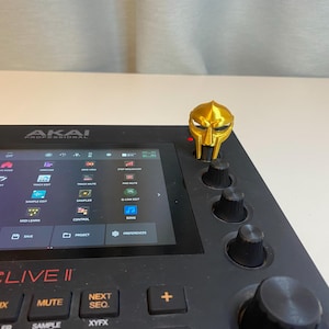 Może przedstawiać: Złoty, wydrukowany w 3D, gałka w kształcie maski 'The RZA' do czarnego samplera do produkcji muzyki Akai MPC Live II.