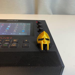 May include: A black Akai MPC Live II music production sampler with a gold-colored plastic mask on top of the device. The screen displays a grid of icons.