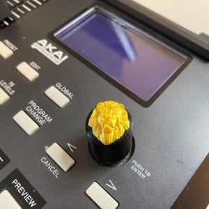 Peut inclure: Gros plan sur un contrôleur de production musicale noir avec un bouton en forme de lion jaune. Le contrôleur comporte des boutons étiquetés "Preset", "Edit", "Global", "16 Levels", "Program Change", "Cancel", "Preview", "Push to Enter", et "Control Bank".