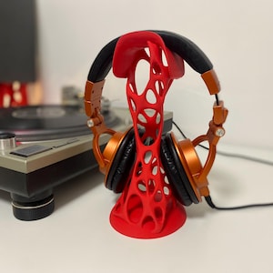 Pode incluir: Suporte vermelho para fones de ouvido com design aberto, segurando fones de ouvido laranja e preto. O suporte está em uma superfície branca, com uma plataforma giratória e um alto-falante ao fundo. Os fones de ouvido têm um cabo preto.