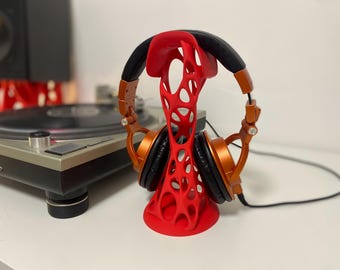 May include: A red, open-design headphone stand holding orange and black headphones. The stand is on a white surface, with a turntable and speaker in the background. The headphones have a black cable.
