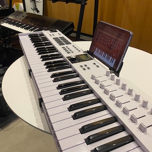 Op de afbeelding: Een wit digitaal keyboard met 61 toetsen, een bedieningspaneel met knoppen en draaiknoppen en een tablet die bovenop is bevestigd. De tablet toont een muziekproductie-app.