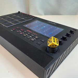 May include: A black Akai MPC Live II music production sampler with a yellow dice on the jog wheel. The screen displays the text "MPC LIVE II".