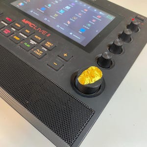 May include: A black MPC Live II music production controller with a touchscreen display, buttons, knobs, and a gold-colored knob with a textured surface.