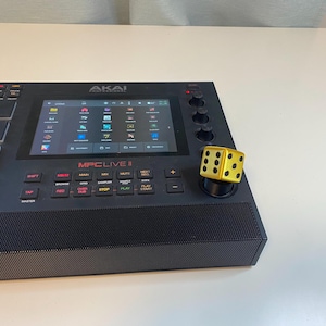 May include: A black Akai MPC Live II music production center with a color display and numerous buttons. A gold-colored die is on the right side. The device is on a white surface.