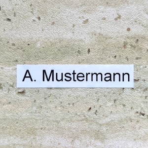 Könnte beinhalten: Ein weißes Etikett mit schwarzem Text, der "A. Mustermann" lautet.