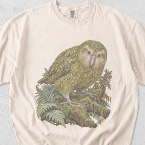 May include: An ivory-coloured short-sleeve t-shirt featuring a detailed illustration of a green kakapo bird perched on a mossy branch surrounded by ferns. The shirt has a crew neckline and a small tag at the collar with the text DD ARTS DESIGN CO.