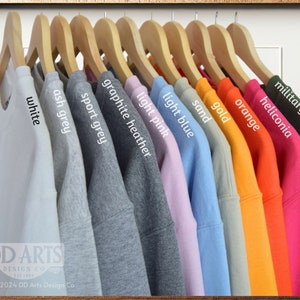 Op de afbeelding: Een rij sweatshirts die aan houten hangers hangen, met een verscheidenheid aan kleuren, waaronder wit, asgrijs, sportgrijs, grafiet melange, lichtroze, lichtblauw, zand, goud, oranje, heliconia en legergroen. De tekst "DD ARTS DESIGN CO" is zichtbaar onderaan de afbeelding.