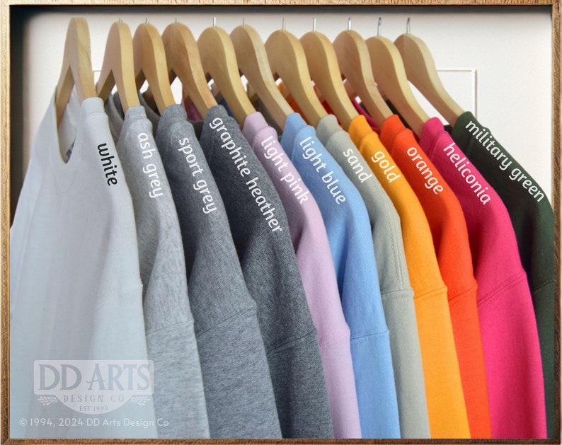 Op de afbeelding: Een rij sweatshirts die aan houten hangers hangen, met een verscheidenheid aan kleuren, waaronder wit, asgrijs, sportgrijs, grafiet melange, lichtroze, lichtblauw, zand, goud, oranje, heliconia en legergroen. De tekst "DD ARTS DESIGN CO" is zichtbaar onderaan de afbeelding.
