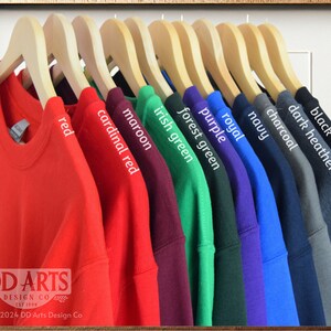 Op de afbeelding: Een rij van tien T-shirts hangen aan houten hangers, met een verscheidenheid aan kleuren, waaronder rood, kardinaalrood, donkerrood, Iers groen, bosgroen, paars, koninklijk blauw, marineblauw, antraciet, donkergrijs gem&ecirc;leerd en zwart. DD Arts Design Co. est. 1994. &copy;1994, 2024 DD Arts Design Co.