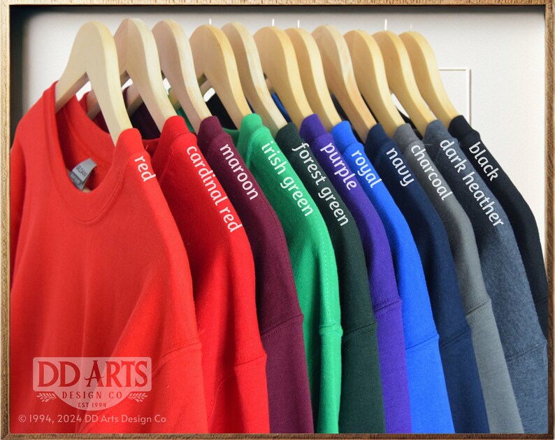 Op de afbeelding: Een rij van tien T-shirts hangen aan houten hangers, met een verscheidenheid aan kleuren, waaronder rood, kardinaalrood, donkerrood, Iers groen, bosgroen, paars, koninklijk blauw, marineblauw, antraciet, donkergrijs gem&ecirc;leerd en zwart. DD Arts Design Co. est. 1994. &copy;1994, 2024 DD Arts Design Co.