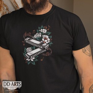 May include: A black t-shirt featuring a graphic design of two harmonicas and white flowers. The harmonicas are silver and black with a floral design. The flowers are white with green leaves. The design is surrounded by musical notes.