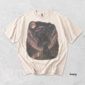 Puede incluir: Camiseta de color marfil con una ilustraci&oacute;n detallada de un Labrador Retriever negro bajo un cielo estrellado con luna llena. El dise&ntilde;o incluye un retrato m&aacute;s grande y una imagen m&aacute;s peque&ntilde;a del perro cerca de un lago y &aacute;rboles.