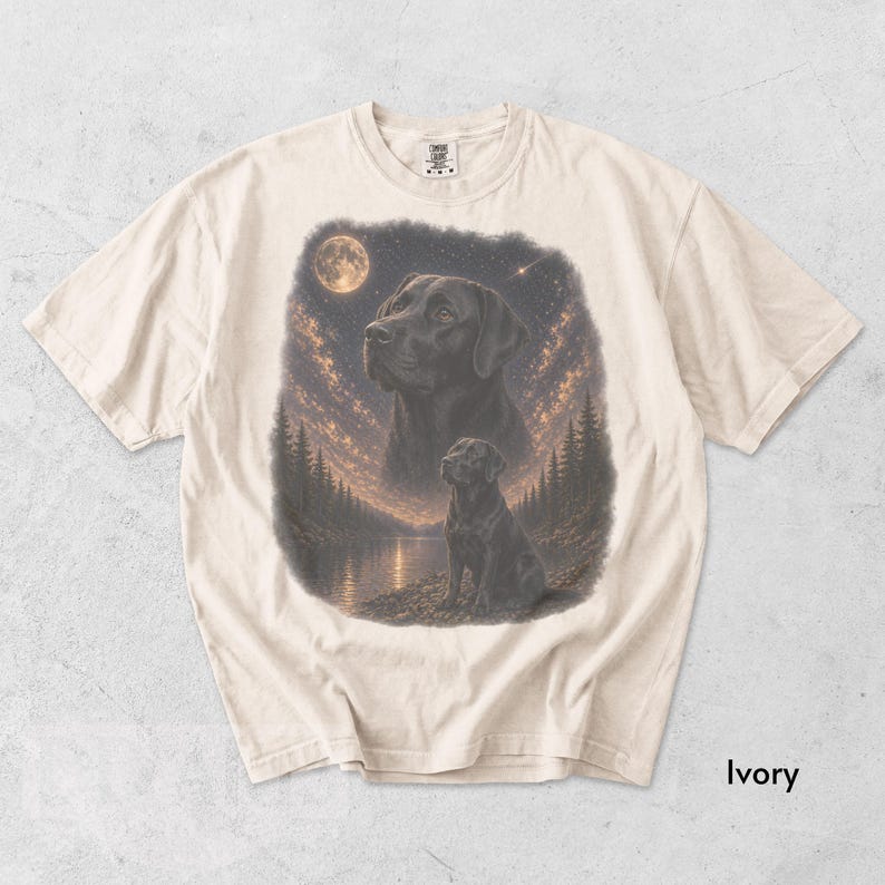 Puede incluir: Camiseta de color marfil con una ilustraci&oacute;n detallada de un Labrador Retriever negro bajo un cielo estrellado con luna llena. El dise&ntilde;o incluye un retrato m&aacute;s grande y una imagen m&aacute;s peque&ntilde;a del perro cerca de un lago y &aacute;rboles.