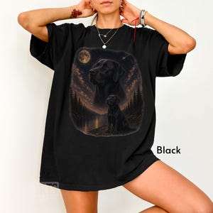 Puede incluir: Camiseta negra con un gr&aacute;fico de un Labrador Retriever negro bajo una luna llena y un cielo estrellado. El dise&ntilde;o incluye un lago y una escena de bosque. La palabra "Black" est&aacute; impresa en la parte inferior derecha.