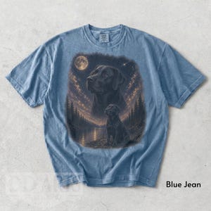 Puede incluir: Camiseta azul vaquero con una ilustraci&oacute;n detallada de un Labrador Retriever negro. La obra de arte incluye un cielo nocturno con luna llena, un lago y pinos. El texto "Blue Jean" est&aacute; impreso en la parte inferior.
