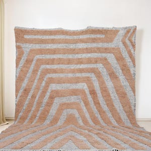 Könnte beinhalten: Ein großer, rechteckiger Teppich mit geometrischem Design. Der Teppich weist ein Muster aus abwechselnden Pfirsich- und grauen Streifen auf, das einen kühnen, modernen Look erzeugt. Der Teppich besteht aus einem weichen, strukturierten Material.